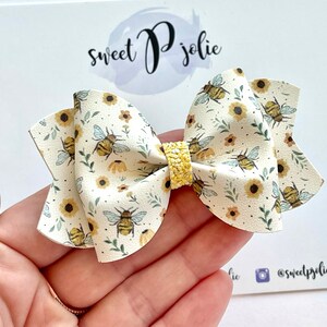 May include: A white fabric bow with a bee and sunflower pattern. The bow has a yellow glitter centre.