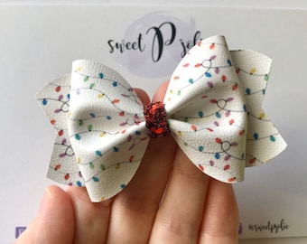 May include: A white bow with a Christmas lights pattern. The bow features a red glitter center and is held in a hand. The bow is a festive accessory, perfect for the holiday season.
