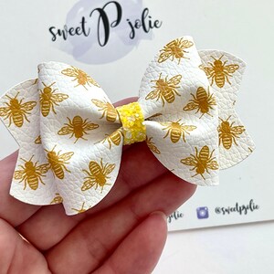 May include: A white bow with a yellow glitter centre and a gold bee pattern. The bow is made of faux leather and has a slightly textured surface.