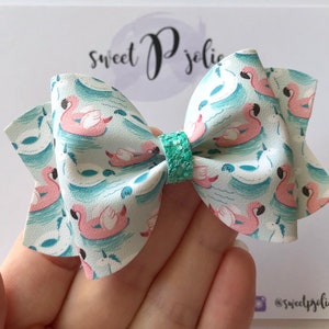 May include: A turquoise and white bow with a pink flamingo print. The bow has a turquoise glitter center.