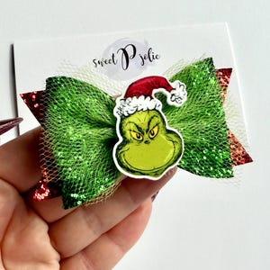 May include: A festive Christmas hair bow in green and red, featuring a Grinch face wearing a Santa hat. The bow is made of glittery green mesh and red ribbon. The bow is held in a hand.