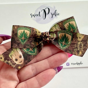 May include: A brown and green bow with a glitter center. The bow is decorated with a camouflage pattern and the words "Guardians" and an image of a character from the movie Guardians of the Galaxy.