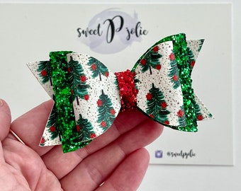 May include: A festive Christmas hair bow featuring a white background with green Christmas trees and red ornaments. The bow has green glitter accents and a red glitter center. The bow is held in a hand. The text "sweet P jolie" is visible.