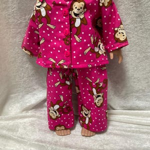 May include: A doll wearing a pink pajama set with a monkey pattern. The pajamas have a button-down top and matching pants. The monkey design is brown and white, with white polka dots on the pink fabric.