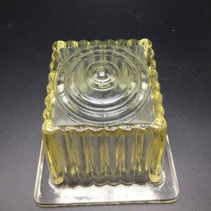 May include: A clear glass square dish with a fluted design and a raised circular center. The dish is made of clear glass and has a yellow tint.