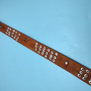 Peut inclure: Une ceinture en cuir marron avec des &oelig;illets en m&eacute;tal argent&eacute;. La ceinture est sur un fond bleu clair.