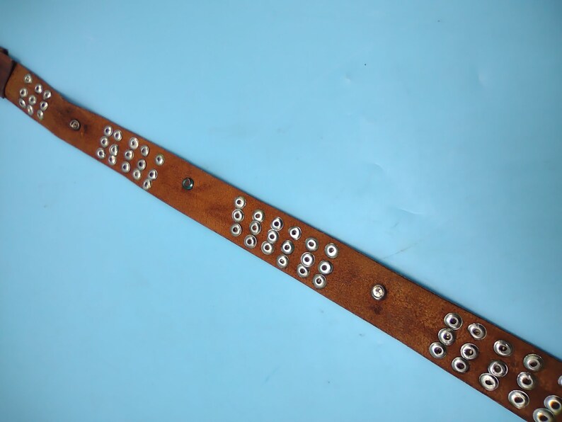 Peut inclure: Une ceinture en cuir marron avec des &oelig;illets en m&eacute;tal argent&eacute;. La ceinture est sur un fond bleu clair.