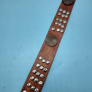 Peut inclure: Une ceinture en cuir marron avec des clous argent&eacute;s et un motif de pi&egrave;ce de monnaie. La ceinture a une rang&eacute;e de clous argent&eacute;s au centre et une pi&egrave;ce de monnaie &agrave; l'extr&eacute;mit&eacute;.