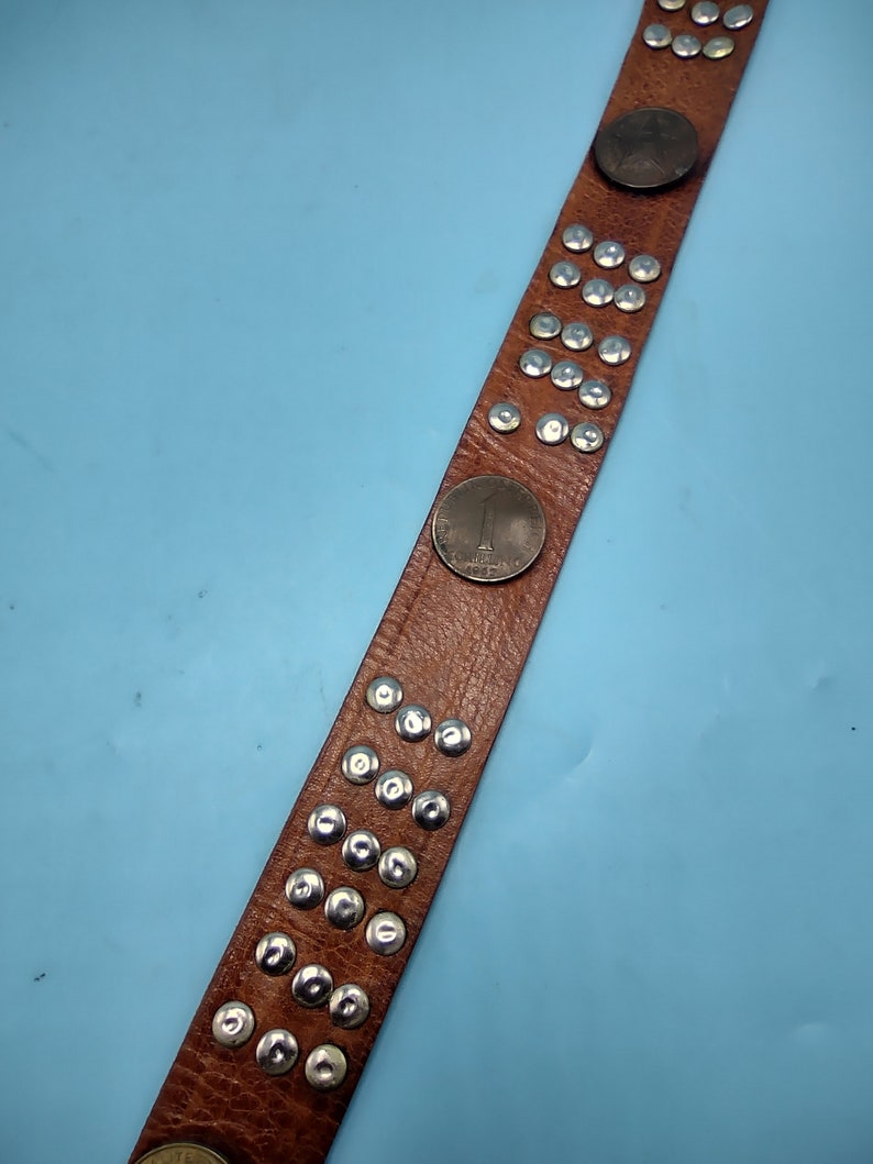 Peut inclure: Une ceinture en cuir marron avec des clous argent&eacute;s et un motif de pi&egrave;ce de monnaie. La ceinture a une rang&eacute;e de clous argent&eacute;s au centre et une pi&egrave;ce de monnaie &agrave; l'extr&eacute;mit&eacute;.