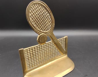 May include: A gold-colored tennis-themed decorative piece. It features a tennis racket, ball, and net, all crafted in a detailed, textured design. The base is a curved shape, and the overall aesthetic is vintage.