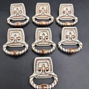 May include: Seven white and gold cabinet pulls with a decorative design. Each pull features a rectangular backplate with a fleur-de-lis motif and a curved handle with a central, cylindrical accent. The pulls are arranged on a dark surface.