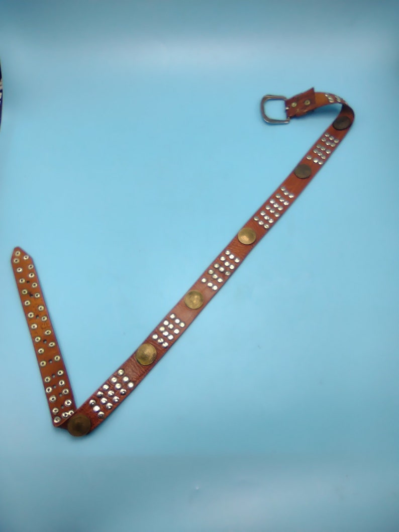 Peut inclure: Une ceinture en cuir marron avec des clous argent&eacute;s et des accents ronds dor&eacute;s. La ceinture a une boucle argent&eacute;e.