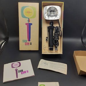 May include: A vintage Sylvania Sun Gun II movie light in its original box. The box is yellow and white with a colorful illustration of the light. The light is silver and black with a clear bulb. The box also includes a pamphlet with instructions on how to use the light.