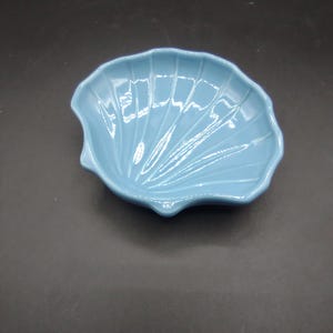 May include: A light blue ceramic dish shaped like a seashell. The dish has a smooth, glossy finish and is perfect for serving small snacks or holding jewelry.