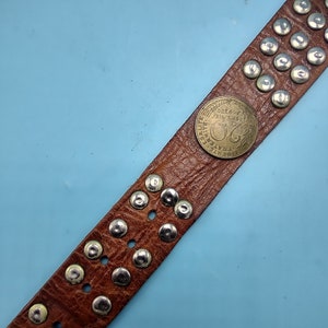 Peut inclure: Une ceinture en cuir marron avec des clous argent&eacute;s et une pi&egrave;ce ronde avec le texte "LIBERTE EGALITE FRATERNITE" et le num&eacute;ro "20" au centre.