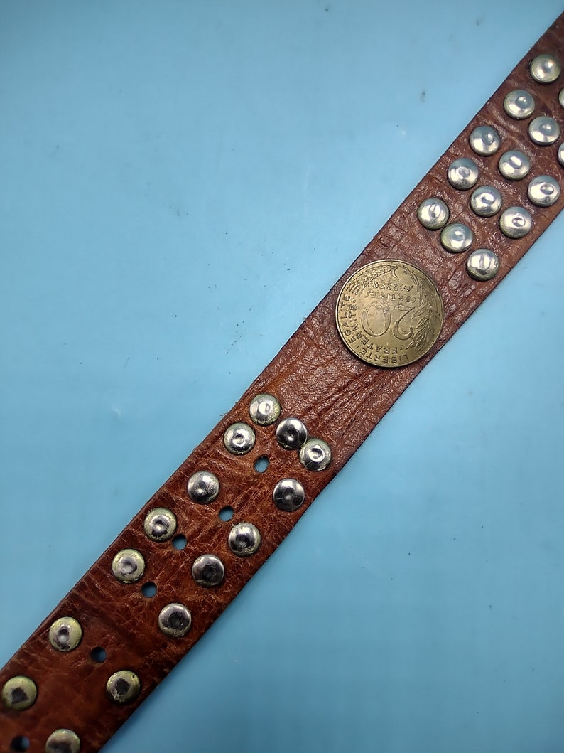 Peut inclure: Une ceinture en cuir marron avec des clous argent&eacute;s et une pi&egrave;ce ronde avec le texte "LIBERTE EGALITE FRATERNITE" et le num&eacute;ro "20" au centre.