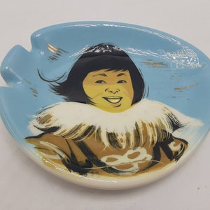 May include: A vintage ashtray with a light blue background and a smiling Inuit child illustration. The child has black hair, a yellow face, and wears a brown coat with white fur trim. Gold accents are present on the ashtray.