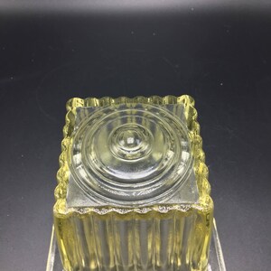 May include: A clear glass square dish with a fluted design and a raised circular center. The dish is made of clear glass and has a yellow tint.