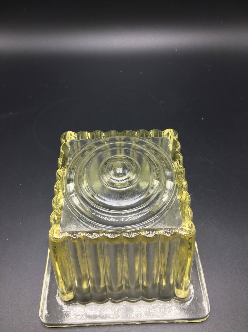 May include: A clear glass square dish with a fluted design and a raised circular center. The dish is made of clear glass and has a yellow tint.