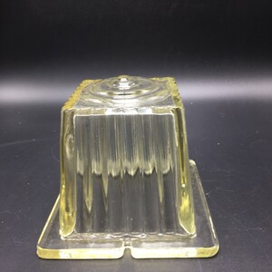 May include: A clear glass square-shaped container with a textured surface and a raised center. The container is made of thick glass and has a slightly yellow tint.