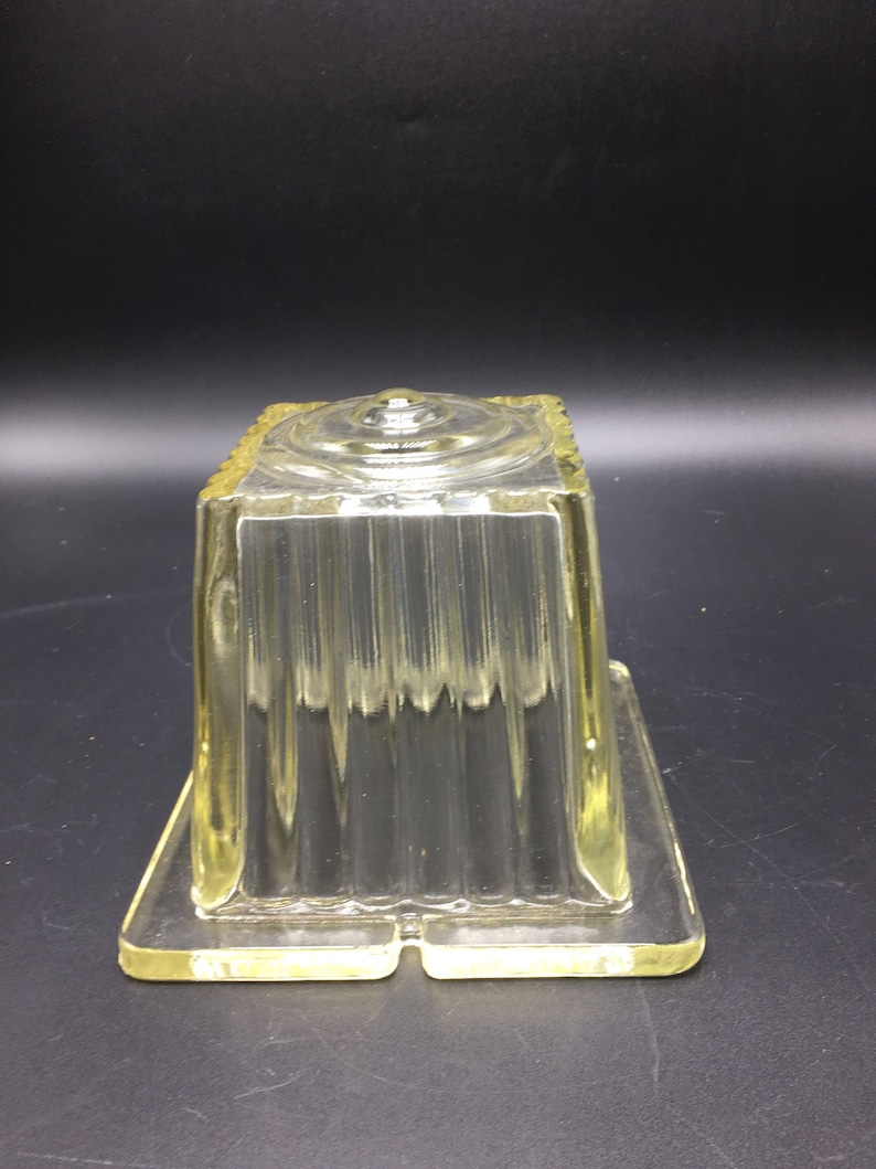 May include: A clear glass square-shaped container with a textured surface and a raised center. The container is made of thick glass and has a slightly yellow tint.