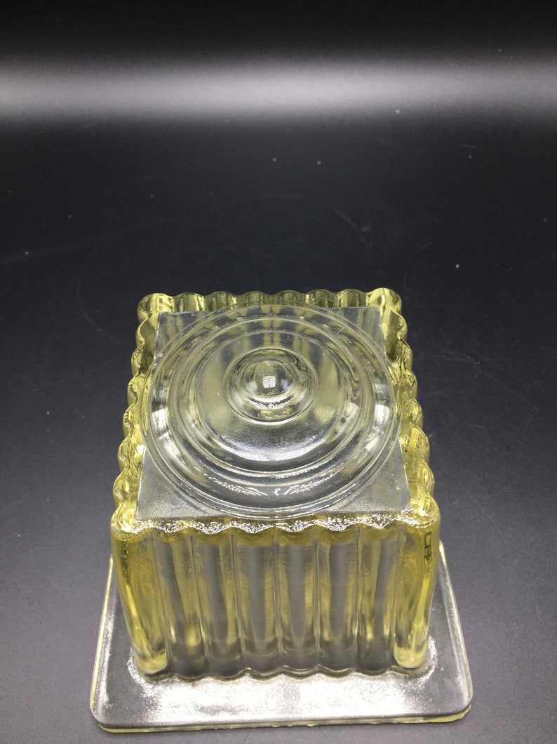 May include: A clear glass square dish with a fluted design and a raised circular center. The dish is made of clear glass and has a yellow tint.