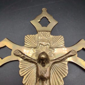K&ouml;nnte beinhalten: Ein goldfarbener Wandkreuz aus Messing mit einer detaillierten Darstellung von Jesus Christus am Kreuz. Das Kreuz hat ein dekoratives Design mit einem Sonnenstrahlenmuster hinter Jesus und den Buchstaben "NR" oben.