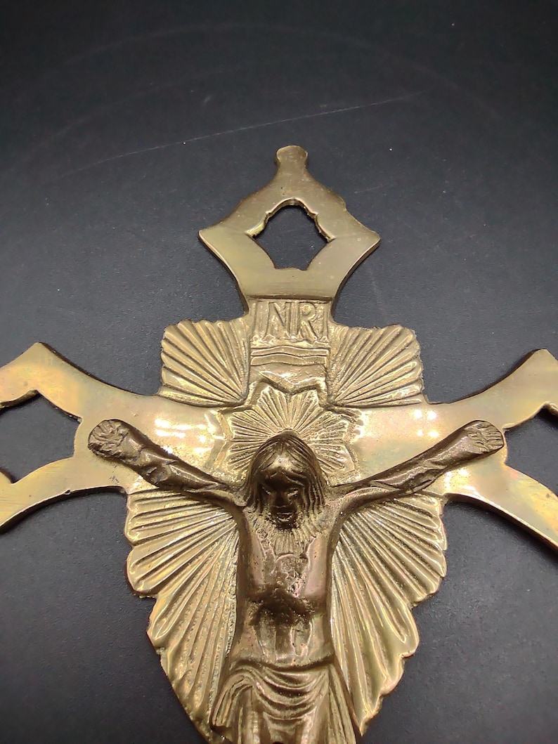 K&ouml;nnte beinhalten: Ein goldfarbener Wandkreuz aus Messing mit einer detaillierten Darstellung von Jesus Christus am Kreuz. Das Kreuz hat ein dekoratives Design mit einem Sonnenstrahlenmuster hinter Jesus und den Buchstaben "NR" oben.