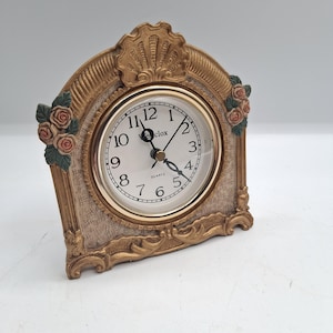May include: A decorative quartz clock with a gold-colored frame. The clock face is white with black numerals and hands. The frame has a textured design with floral accents and a shell-like top.