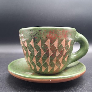 May include: Green ceramic teacup and saucer set. The cup features a geometric diamond pattern in a lighter brown color, with a green handle. The saucer has a matching green color with a brown rim. The cup has some text on the side.