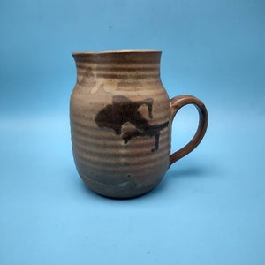 May include: A brown ceramic pitcher with a handle and a dark brown design. The pitcher has a textured surface and a rounded shape.