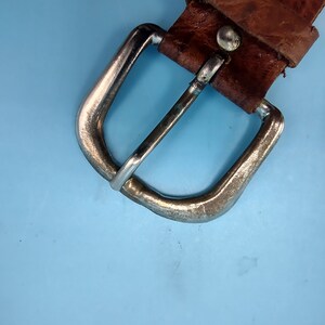 Peut inclure: Gros plan sur une ceinture en cuir marron avec une boucle argent&eacute;e. La boucle est de forme rectangulaire simple avec des bords arrondis.