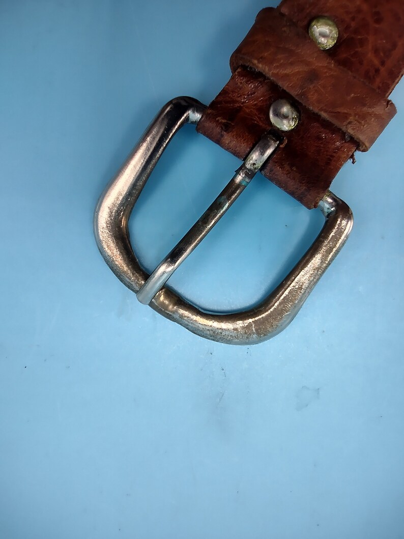 Peut inclure: Gros plan sur une ceinture en cuir marron avec une boucle argent&eacute;e. La boucle est de forme rectangulaire simple avec des bords arrondis.