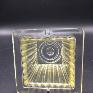 May include: A clear glass ashtray with a square shape and a ribbed interior. The ashtray has a small opening on one side for disposing of cigarette ashes.