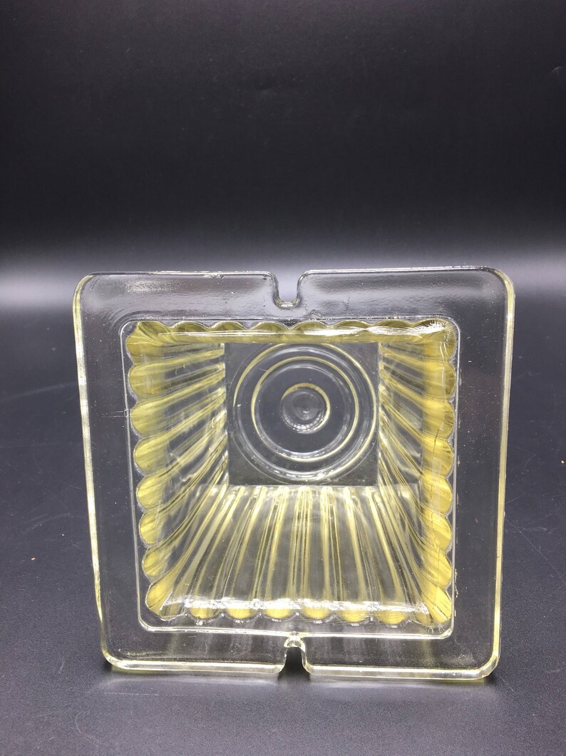 May include: A clear glass ashtray with a square shape and a ribbed interior. The ashtray has a small opening on one side for disposing of cigarette ashes.