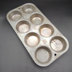 May include: A vintage metal muffin tin with seven individual cups. The tin is silver with some rust spots. The tin has the words "Made in America" printed on the bottom.