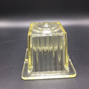 May include: A clear glass butter dish with a square shape and a ribbed design. The dish has a lid with a circular knob on top.