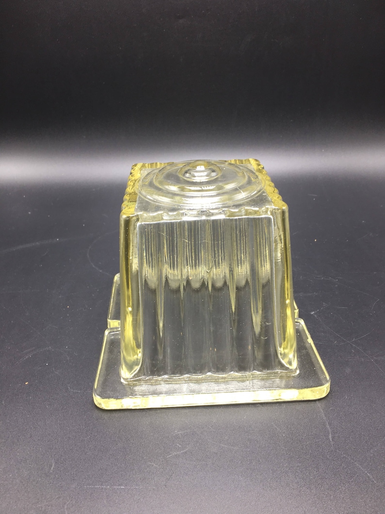May include: A clear glass butter dish with a square shape and a ribbed design. The dish has a lid with a circular knob on top.