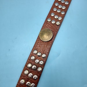 Peut inclure: Une ceinture en cuir marron avec des clous argent&eacute;s et un accent de pi&egrave;ce de monnaie en laiton. La ceinture est sur un fond bleu clair.
