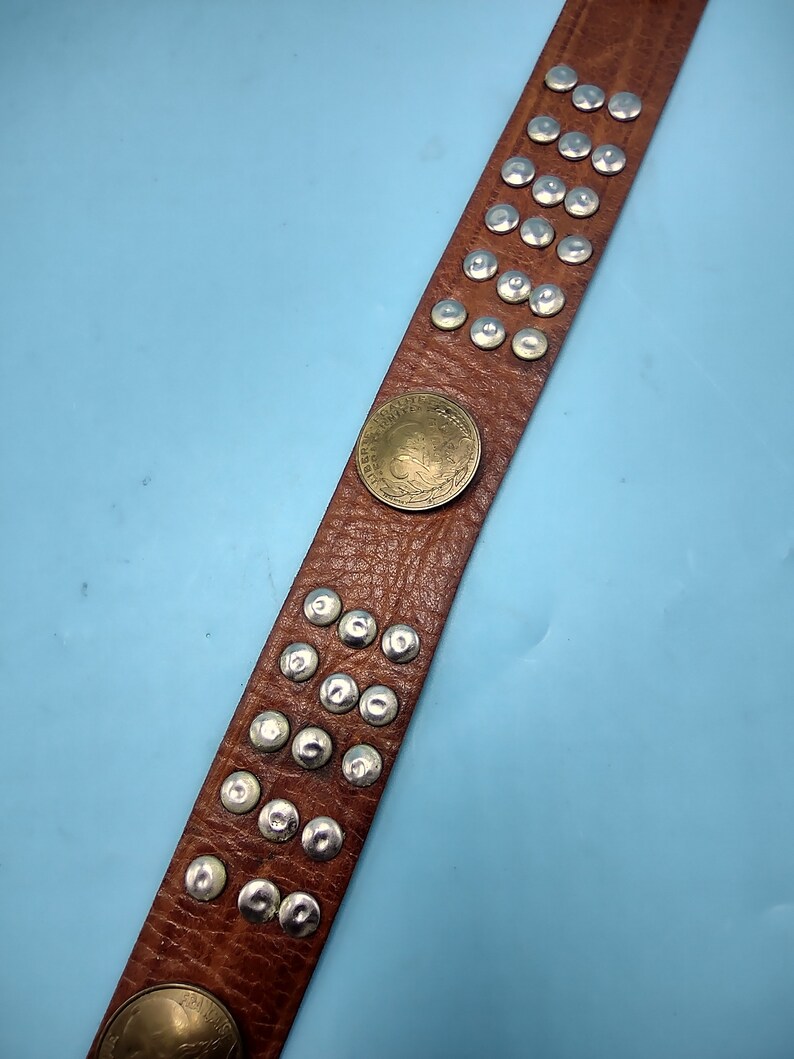 Peut inclure: Une ceinture en cuir marron avec des clous argent&eacute;s et un accent de pi&egrave;ce de monnaie en laiton. La ceinture est sur un fond bleu clair.
