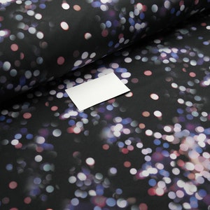 Può includere: Tessuto nero con un motivo di cerchi colorati sfocati nelle tonalità del bianco, rosa e blu. Una carta rettangolare bianca è appoggiata sul tessuto piegato. Il tessuto ha un effetto bokeh.