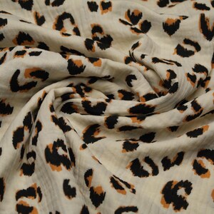 Könnte beinhalten: Cremefarbener Stoff mit einem sich wiederholenden Leopardenmuster. Das Muster zeigt schwarze und orangebraune Flecken, die über den Stoff verstreut sind. Der Stoff ist gerafft und gefaltet, wodurch die Textur und der Fall des Materials sichtbar werden.