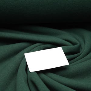 Op de afbeelding: Close-up van donkergroene stof met een zachte textuur, in plooien gelegd. Een blanco witte rechthoekige kaart ligt op de stof. De stof lijkt een rol te zijn.
