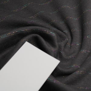 Può includere: Tessuto nero con sottili linee ondulate di glitter iridescente. Il tessuto è drappeggiato su una superficie bianca.