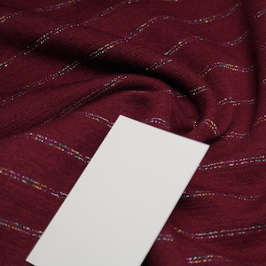 Può includere: Primo piano di un tessuto color borgogna con sottili strisce iridescenti. Un'etichetta bianca vuota è posizionata sul tessuto.