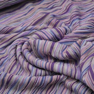 Pode incluir: Close-up de um tecido com um padrão listrado em tons de roxo, rosa, azul e cinza. O tecido é macio e texturizado, com uma aparência ligeiramente ondulada.