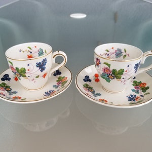 May include: Two white teacups and saucers with a floral pattern in shades of blue, green, orange, and red. The cups have gold trim around the rim.