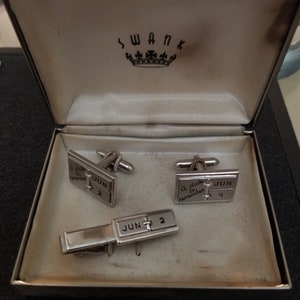 May include: A silver cufflink set with a date and the words "A date to remember" on each cufflink. The set also includes a tie clip with the same date. The cufflinks and tie clip are in a white box with the word "SWANK" printed on the lid.