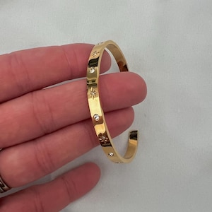 May include: A gold-toned metal cuff bracelet featuring a series of alternating star-shaped engravings and small, clear, round inset stones along the outer surface. The bracelet has an open-ended design with a smooth, polished finish throughout.