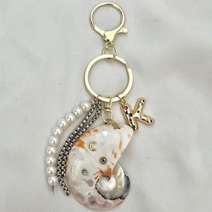 May include: A gold-coloured keyring with a seashell pendant, pearl and chain accents, and a jewelled letter "K" charm. The seashell is white, tan, and grey with a pearlescent sheen. The keyring has a lobster clasp and a split ring.